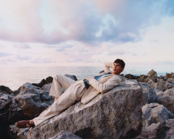 Fashionable man in a sand-colored suit reclining on rocks by the sea. Blue shirt peeks from jacket in outdoor coastal setting.