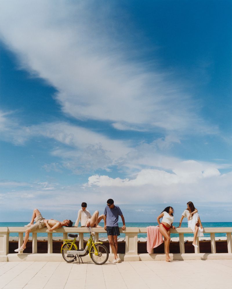 Group of five young adults pose near a yellow moped on a concrete barrier by the sea against a blue sky with clouds on a sunny day.