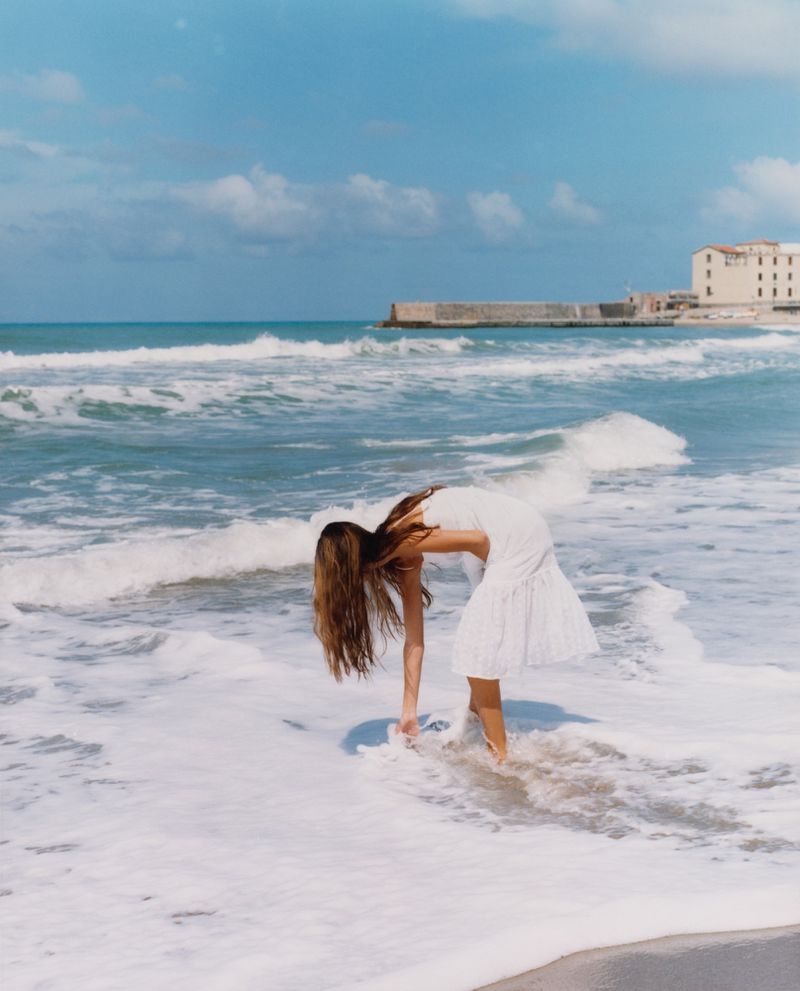 Woman in white dress reaches into the surf on a sunny beach with a building in the distance and a blue sky with scattered clouds.