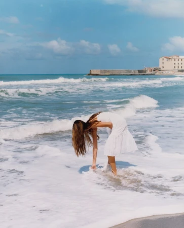 Woman in white dress reaches into the surf on a sunny beach with a building in the distance and a blue sky with scattered clouds.