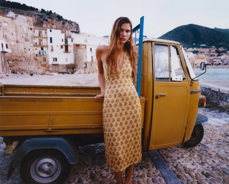 A woman stands next to a yellow truck in a coastal town; she is wearing a patterned dress with a city and ocean in the background.