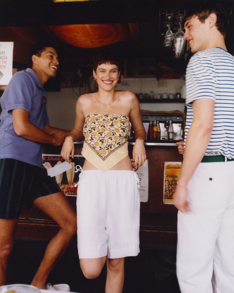 Three adults smile outdoors: a woman in a patterned scarf top, a man in polo shirt, and a man in a striped shirt and white pants.