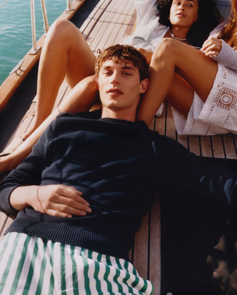 Man lying on the wooden deck of a boat near two seated women, wearing casual clothing with an ocean background on a sunny day.