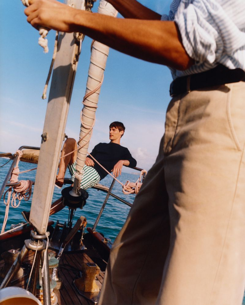 Two young adult men on a sailboat at sea under a blue sky. One is seated wearing striped shorts; another stands in front wearing beige trousers.
