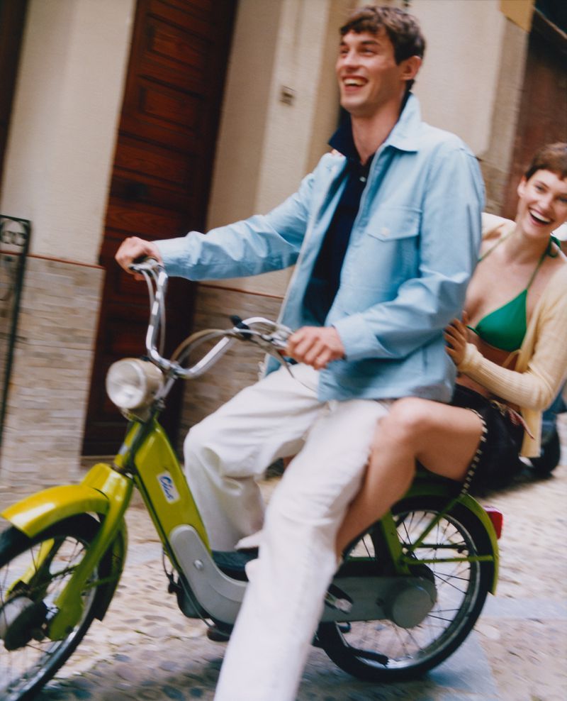 Smiling man and woman ride a lime green scooter down a cobblestone street with buildings on either side on a sunny day.
