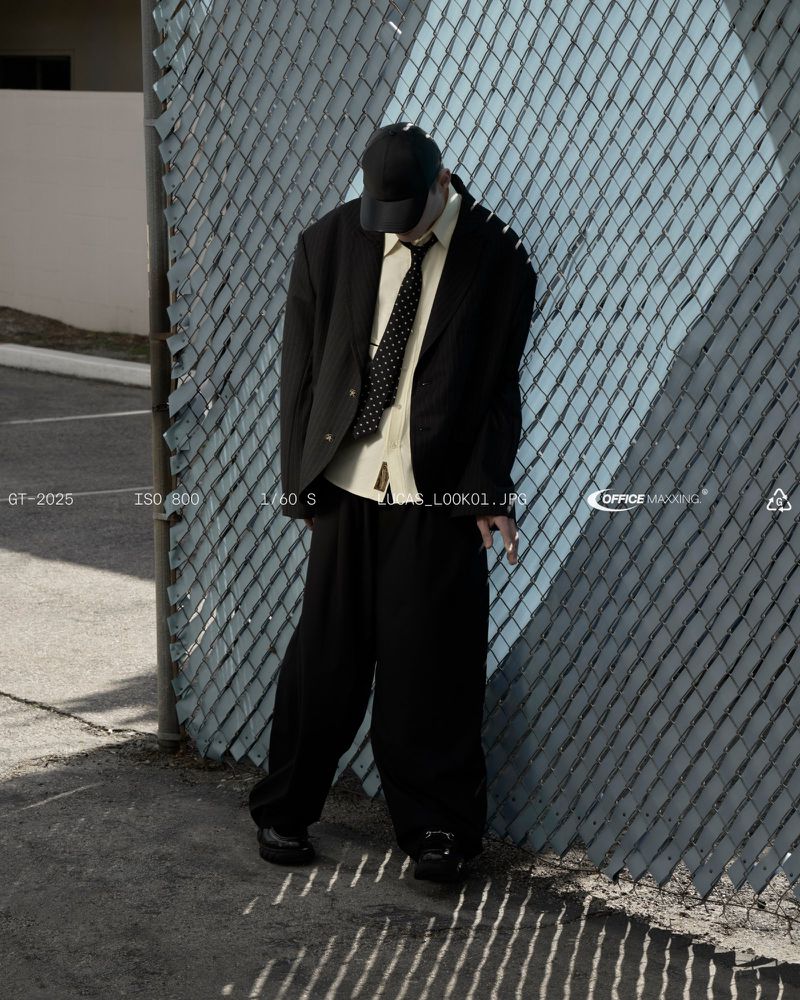 A person in an oversized black suit and baseball cap stands against a blue chain-link fence, shadows adding texture and depth to the shot.