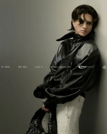 Adult female poses in a black jacket and white pants indoors, leaning against a wall in a studio setting. Fashion photography.