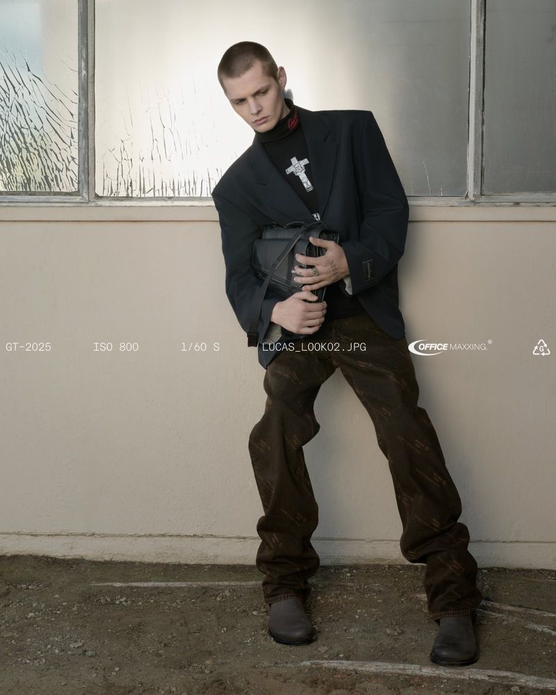 Man poses in a blazer, dark turtleneck with cross pendant, patterned pants, and black bag against an industrial wall with a window.
