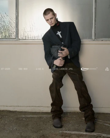 Man poses in a blazer, dark turtleneck with cross pendant, patterned pants, and black bag against an industrial wall with a window.