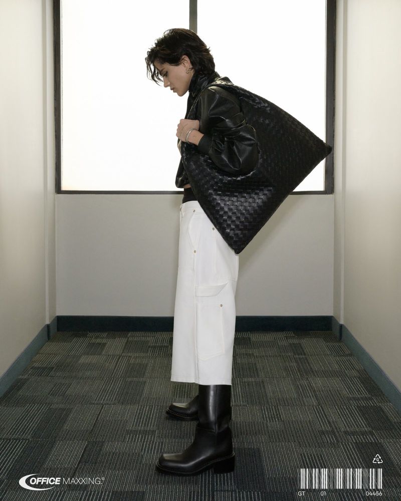 Young adult carries woven black bag over shoulder in interior setting with white walls, wearing a black jacket, white pants and black boots.
