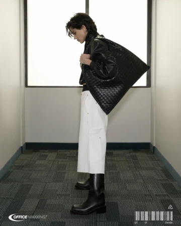 Young adult carries woven black bag over shoulder in interior setting with white walls, wearing a black jacket, white pants and black boots.