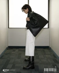 Model in black leather jacket holding large woven bag and wearing white trousers in office corridor