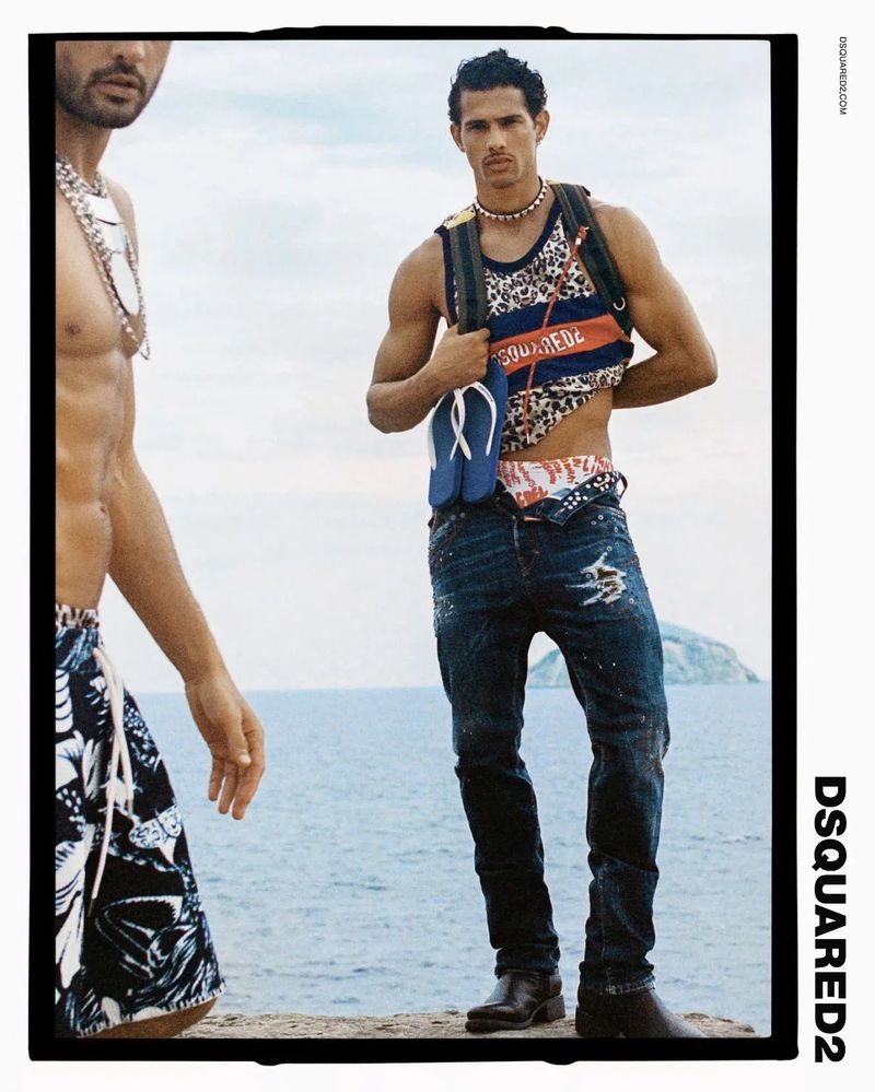 Fashion editorial shot featuring two athletic adult men wearing casual summer clothes and jewelry with a bright, outdoor ocean backdrop.