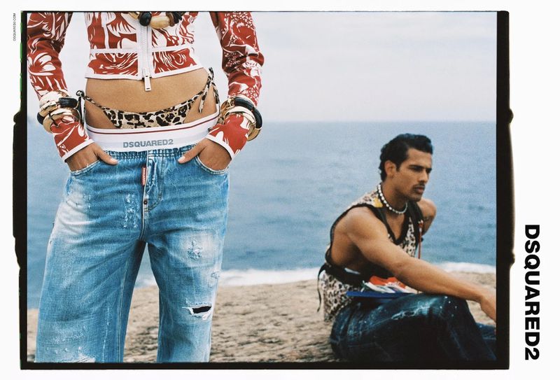 Fashion shot of two models wearing DSQUARED2 clothing, posing on a coastal cliff with a blue sea backdrop and clear sky.