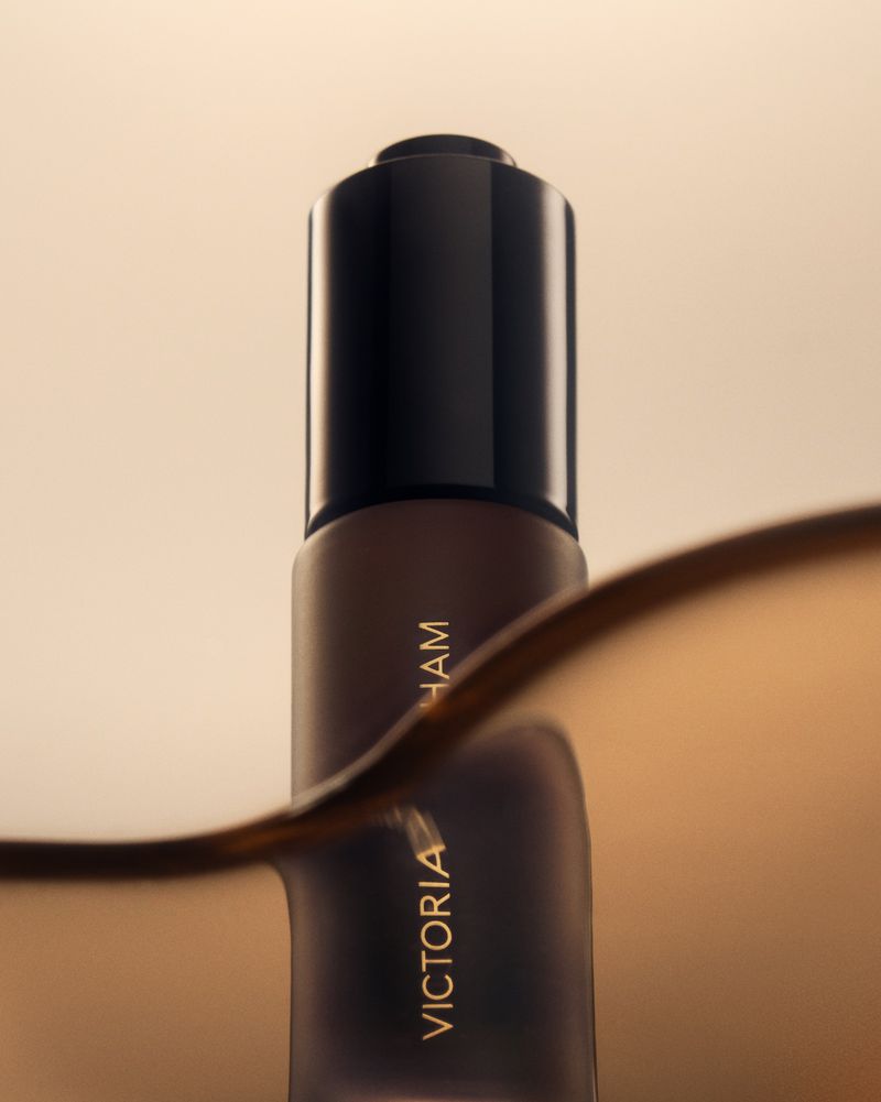 Close-up product shot of a brown serum bottle featuring black cap with gold text against a tan background with a wave effect.