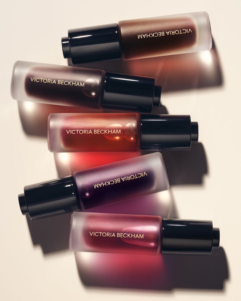 Stacked bottles of Victoria Beckham beauty product in varying shades of color with black caps in studio lighting on plain background.
