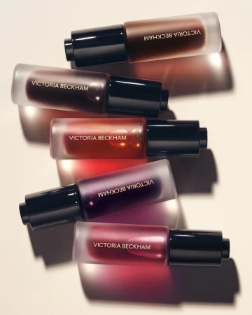 Stacked bottles of Victoria Beckham beauty product in varying shades of color with black caps in studio lighting on plain background.