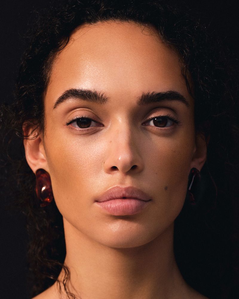 Close-up portrait of an adult woman with dark curly hair and brown eyes, wearing statement earrings against a black background.
