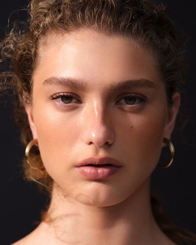 Close-up portrait of a young adult woman with curly hair and gold hoop earrings against a dark background.
