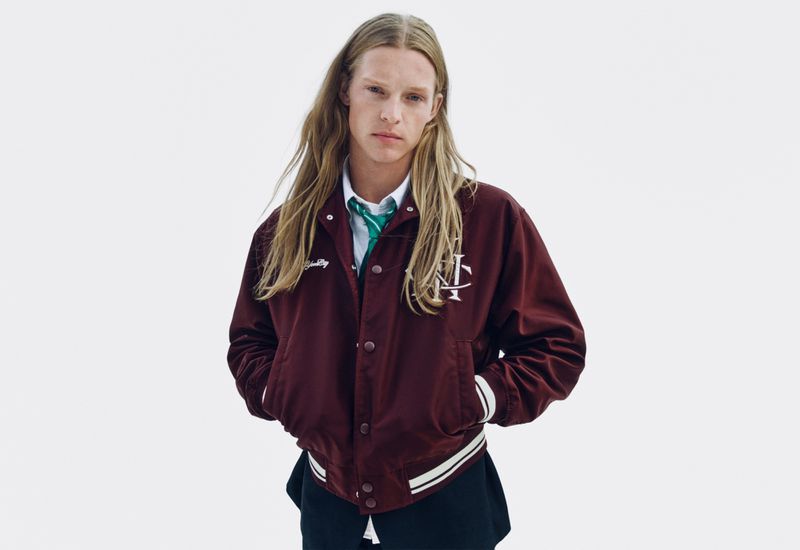 Model with long blonde hair wearing a burgundy varsity jacket over a white shirt and green tie against a white background