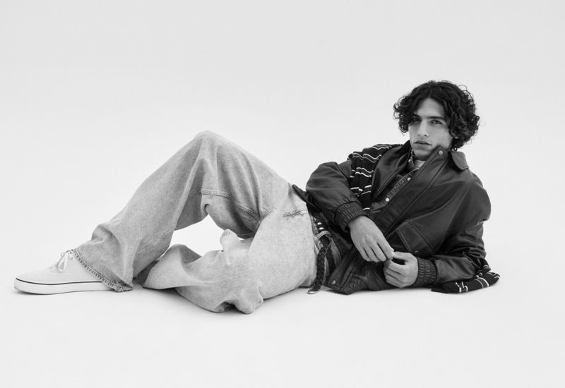 Black-and-white studio shot of a young adult male in an oversized leather jacket and jeans, lying down and looking at the camera.