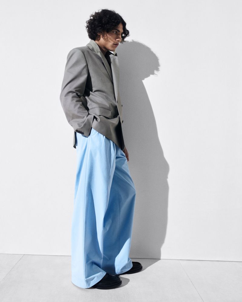 A young male model with dark curly hair wearing a grey blazer and light blue trousers against a plain white studio background.