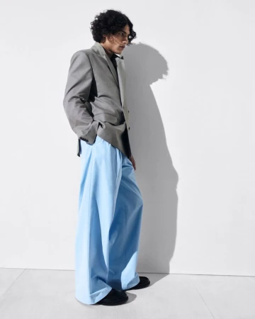 A young male model with dark curly hair wearing a grey blazer and light blue trousers against a plain white studio background.