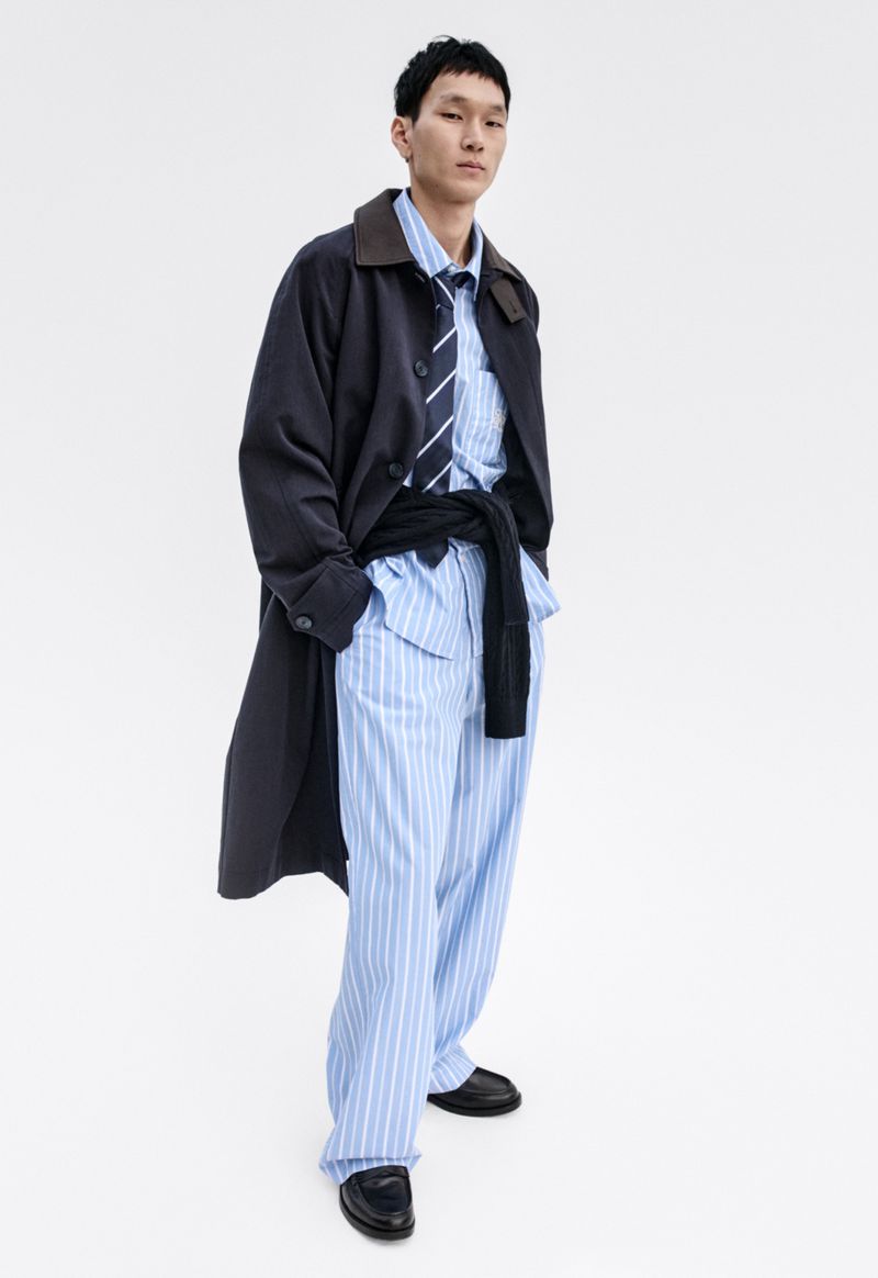 Asian male fashion model poses in a striped pajama set with tie and black trench coat in a studio setting with soft, diffused lighting.