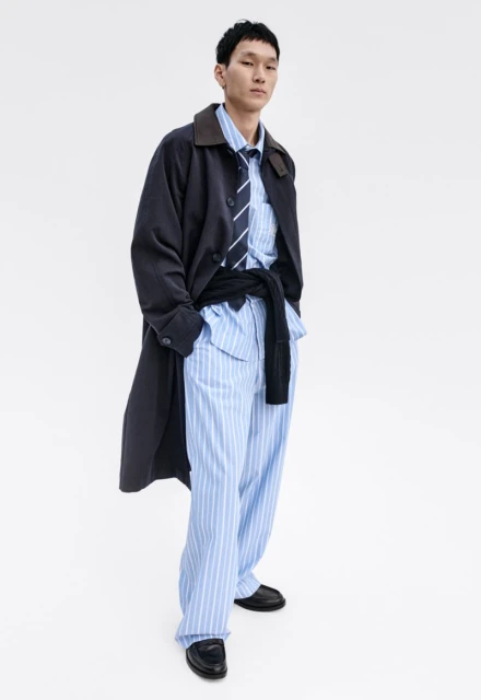 Asian male fashion model poses in a striped pajama set with tie and black trench coat in a studio setting with soft, diffused lighting.