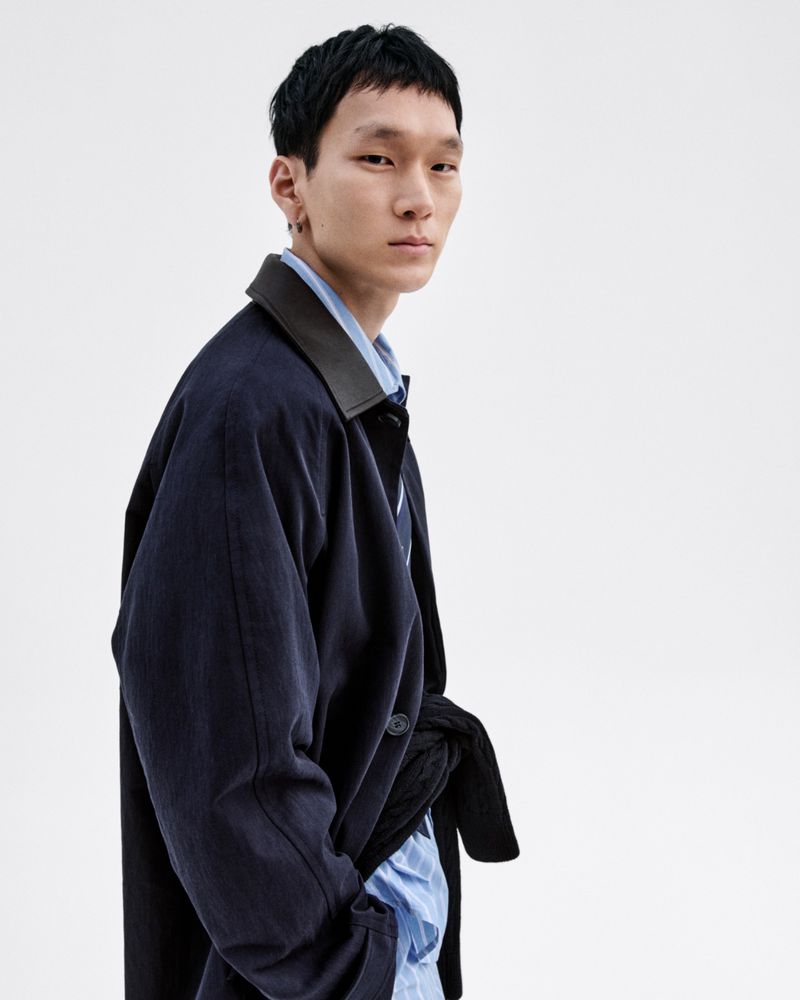 An Asian male model poses in a navy corduroy jacket with a black collar and a light blue shirt against a bright white backdrop in a studio.