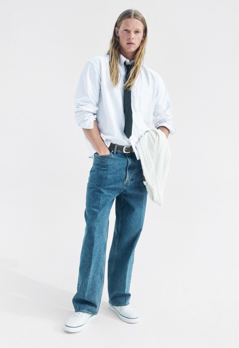 Full-body shot of a person with long blond hair posing in a white collared shirt, blue jeans, white sneakers, and a dark-colored tie.