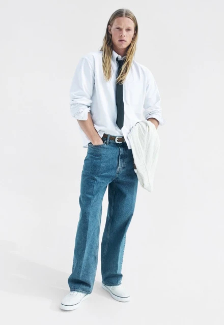 Full-body shot of a person with long blond hair posing in a white collared shirt, blue jeans, white sneakers, and a dark-colored tie.