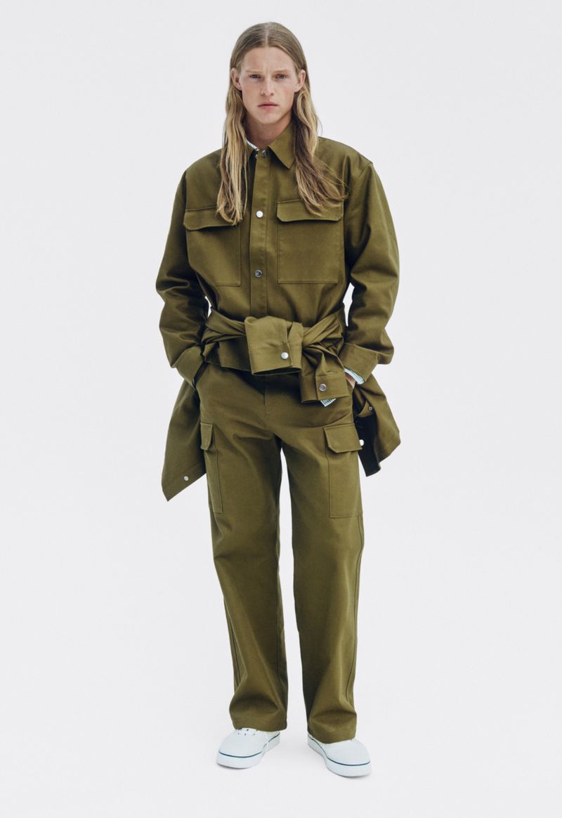 Full-length studio shot of a young adult male fashion model in a green two-piece pantsuit and white sneakers standing against a white backdrop.