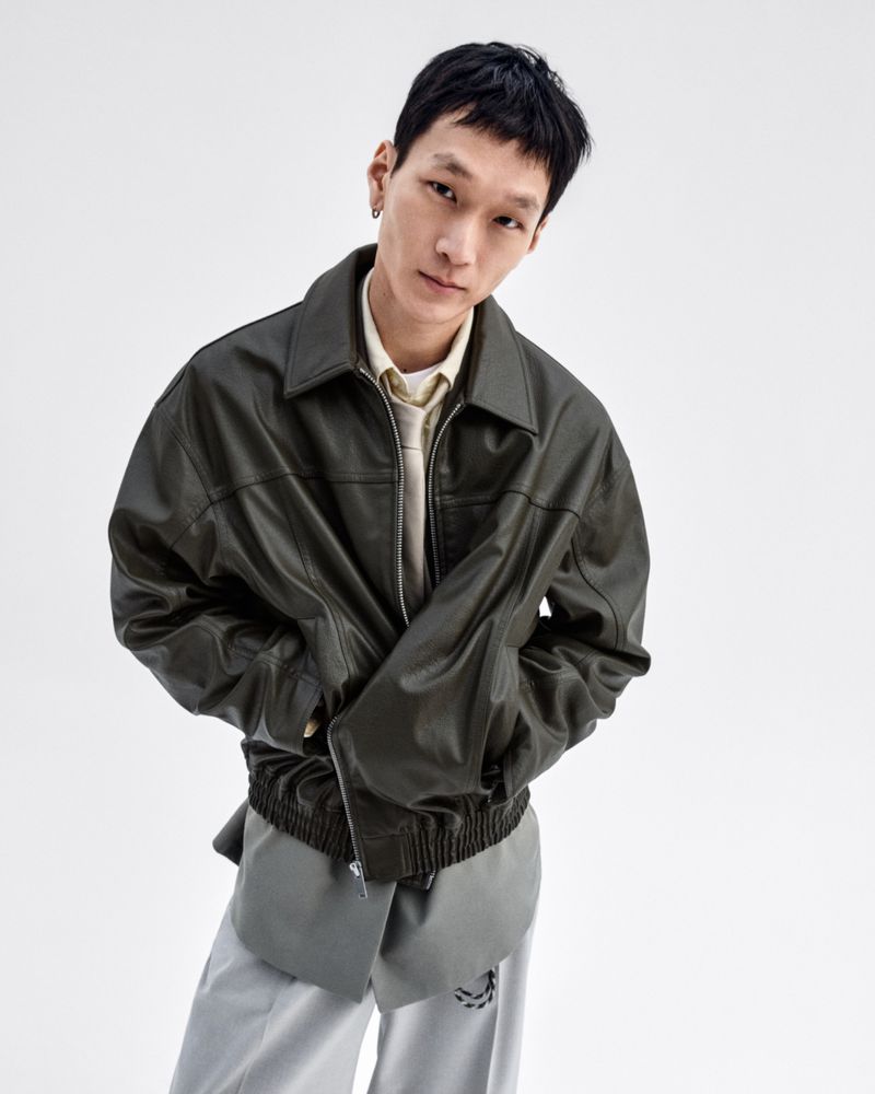 Portrait of an Asian adult male wearing a leather jacket and grey pants, posing in a photo studio setting, against a clean white backdrop.