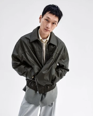 Portrait of an Asian adult male wearing a leather jacket and grey pants, posing in a photo studio setting, against a clean white backdrop.