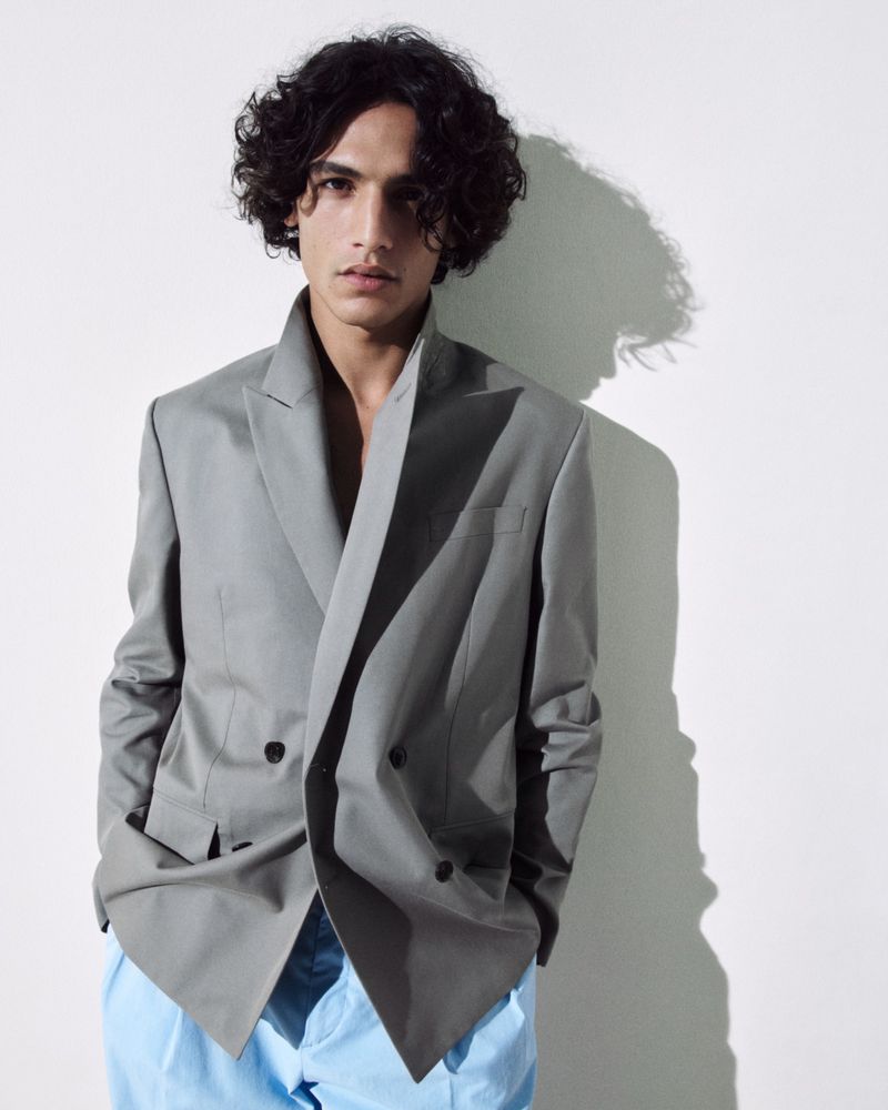 A young adult male with curly hair posing in a gray blazer and blue pants against a white backdrop, lit by studio lighting.