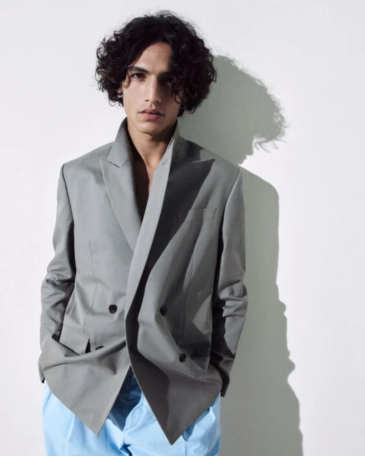 A young adult male with curly hair posing in a gray blazer and blue pants against a white backdrop, lit by studio lighting.