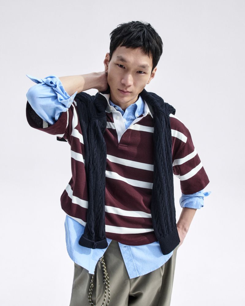 Portrait of a young adult man in a striped maroon rugby shirt and a blue button-down shirt, with a dark blue sweater draped around his neck.