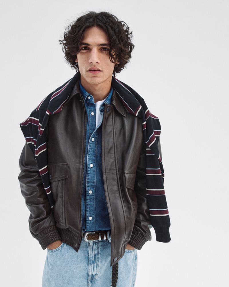 Portrait of a young adult male with curly hair, wearing a brown leather jacket and light blue jeans against a white background.