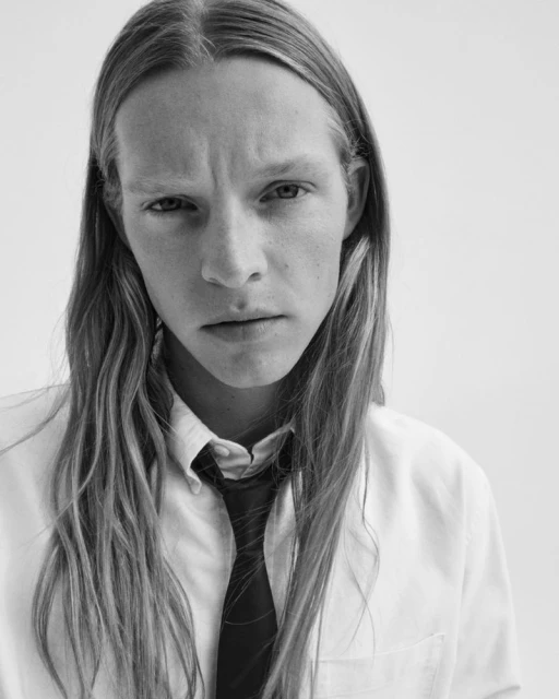 Monochrome portrait of a young adult male with long blond hair wearing a white button-down shirt and a dark tie, gazing directly at the camera.