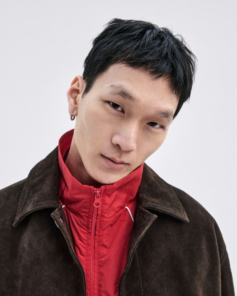 Portrait of an Asian man in a dark brown jacket and red collared shirt against a white background, gazing at the camera with a slight smile.