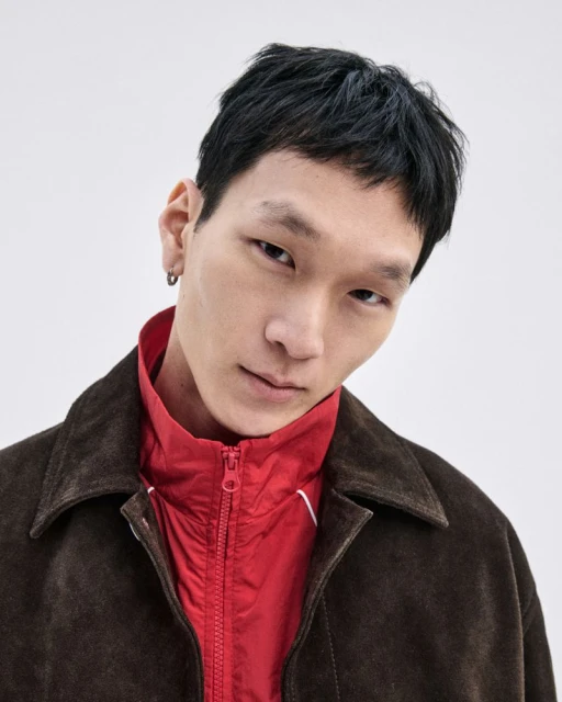 Portrait of an Asian man in a dark brown jacket and red collared shirt against a white background, gazing at the camera with a slight smile.