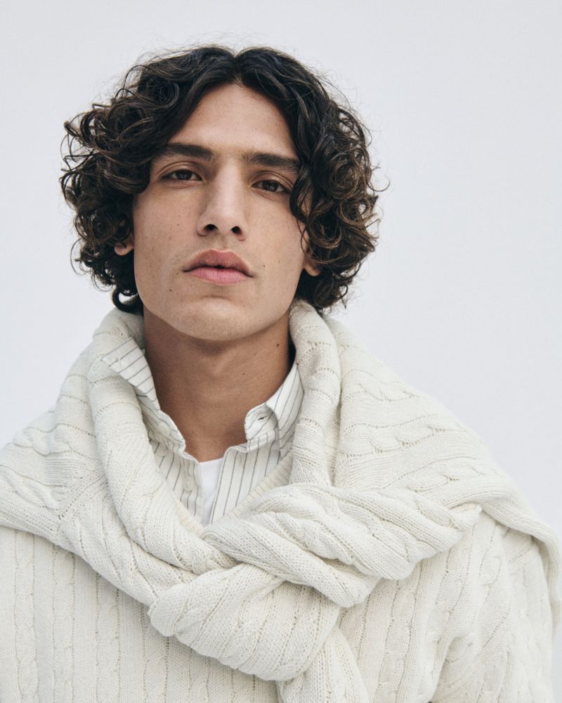 Portrait of a young adult man with curly hair and a knitted sweater draped over his shoulders in a clean, eye-level studio shot.