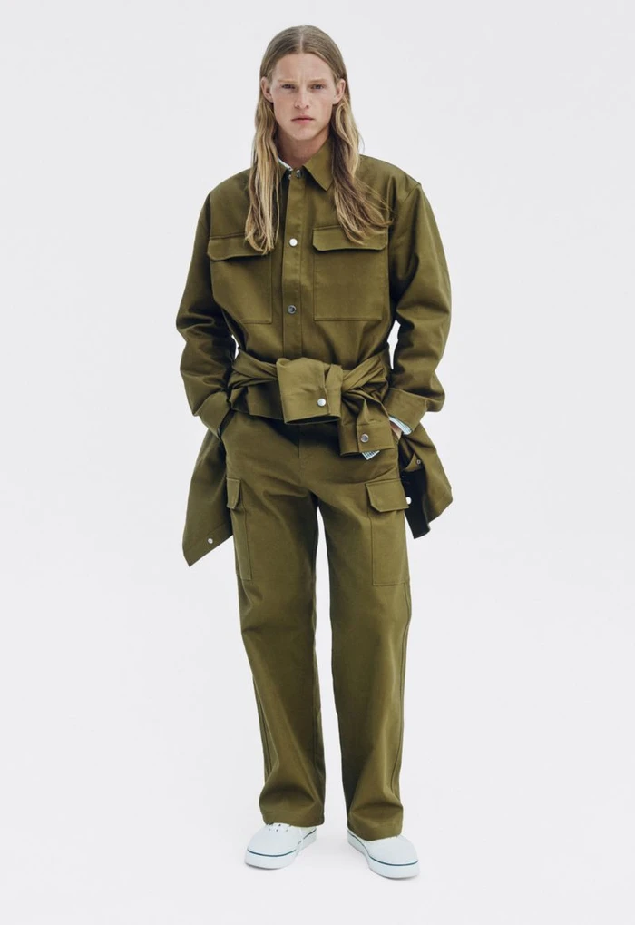 Male model with long blonde hair wearing olive green utilitarian shirt and cargo trousers against a white backdrop.