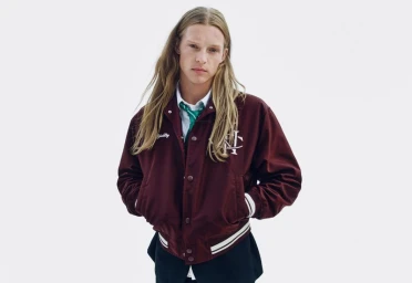 Model with long blonde hair wearing a burgundy varsity jacket over a white shirt and green tie against a white background