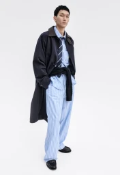 Model wearing a dark coat over striped shirt and trousers against a white background for H&M man spring campaign 2026