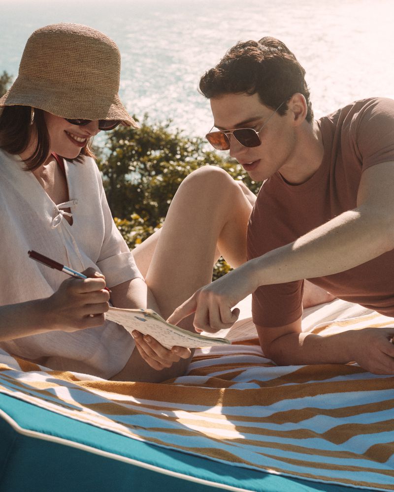 A couple on a striped blanket overlooking the ocean. One person is pointing to a notebook while the other writes with a pen.