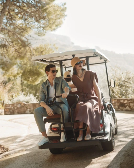 Three people ride in a golf cart on a paved road surrounded by trees. A man drives, a woman sits beside him, and a third person sits in back.
