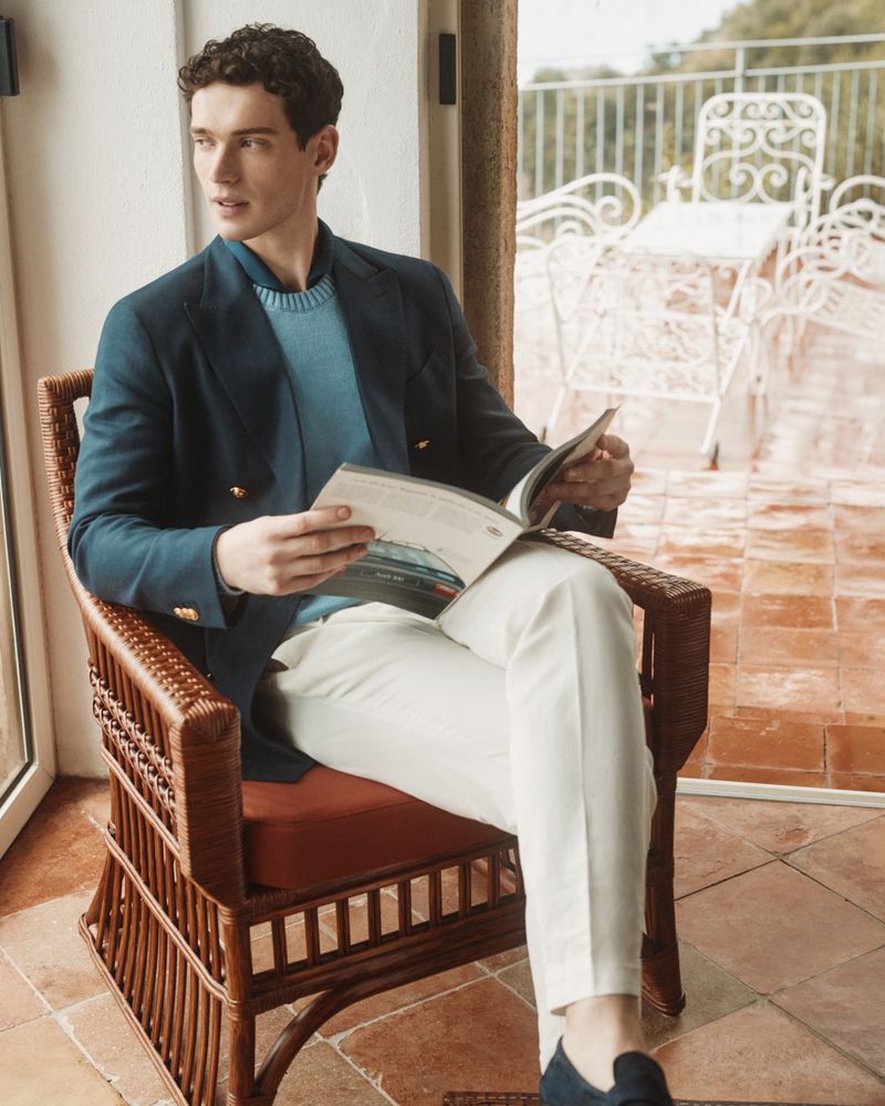 Man in blazer sits in wicker chair reading magazine on tile balcony, outdoor patio furniture in background, neutral and blue tones.