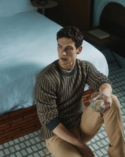 Indoor shot of a brunette man seated on a floor in a sweater and pants, holding a clear glass in a neutral setting.